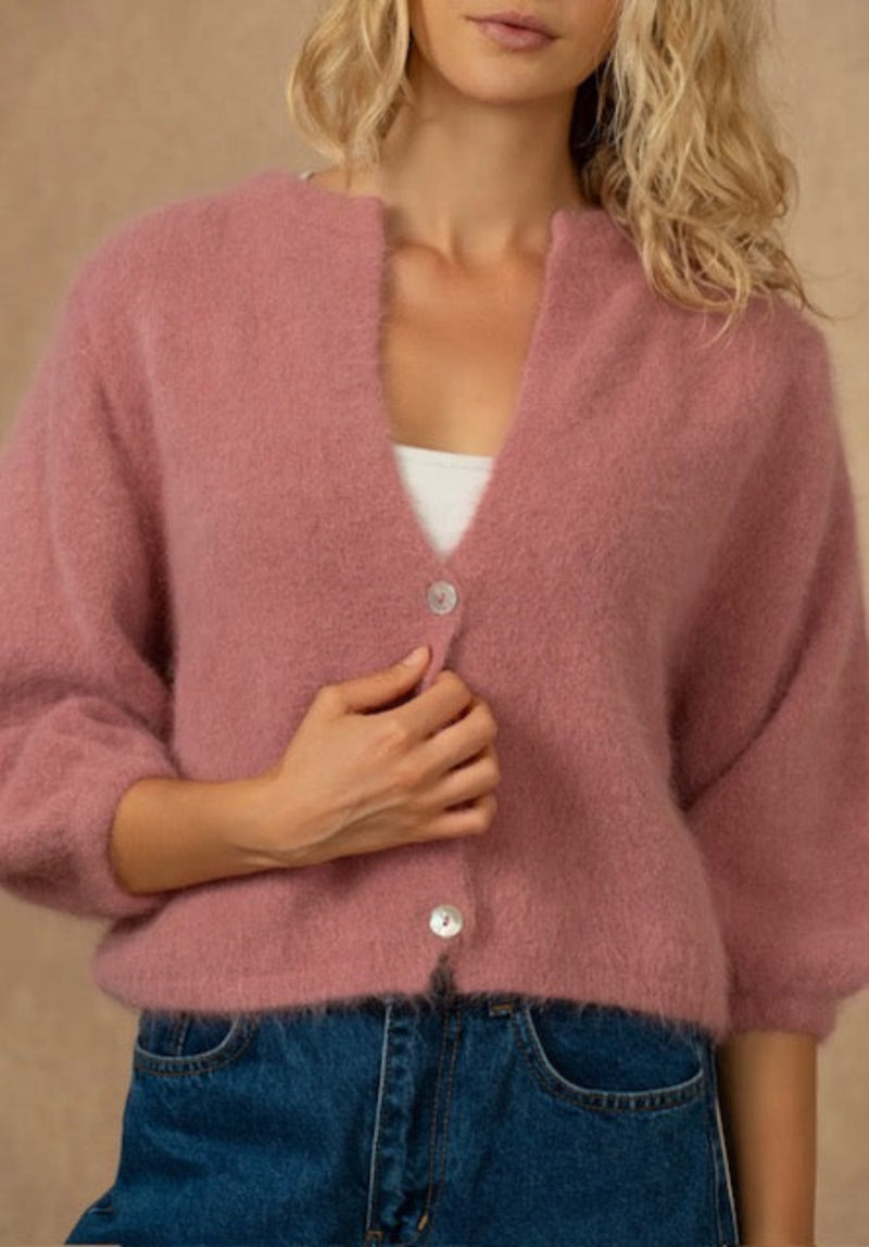 A woman wearing a fuzzy dusty pink cardigan with silver buttons over a white top and blue jeans, set against a warm beige background.