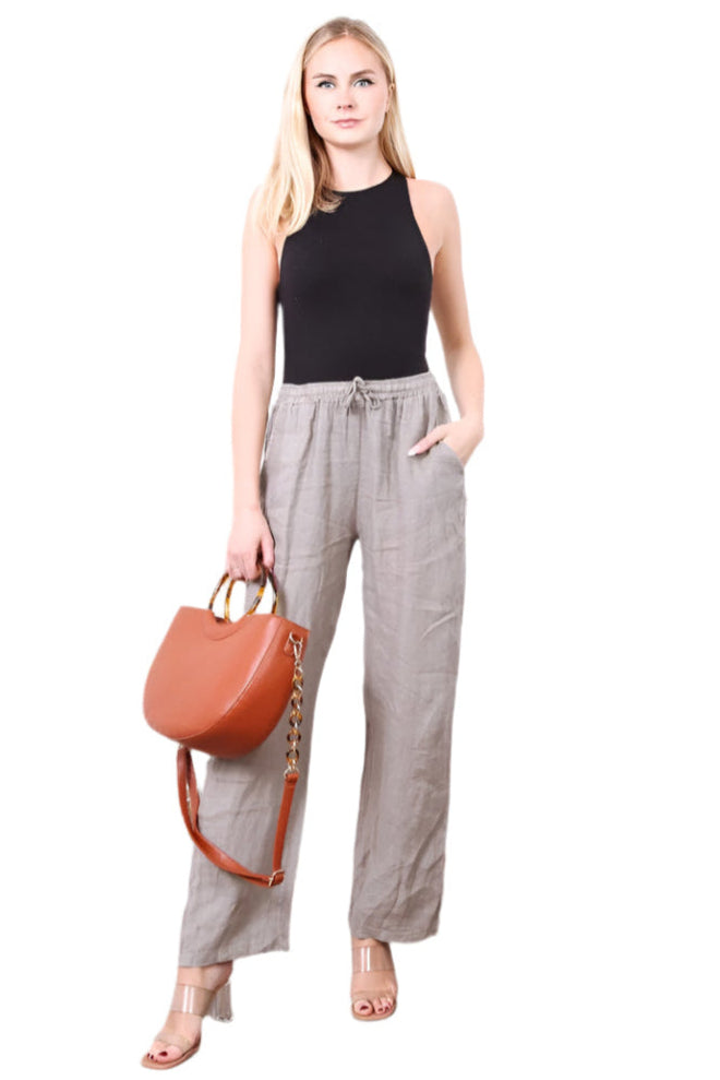 A woman standing against a white background wearing straight leg taupe trousers with an elasticated waist, paired with a black sleeveless top. She is also carrying a handbag and wearing sandals.