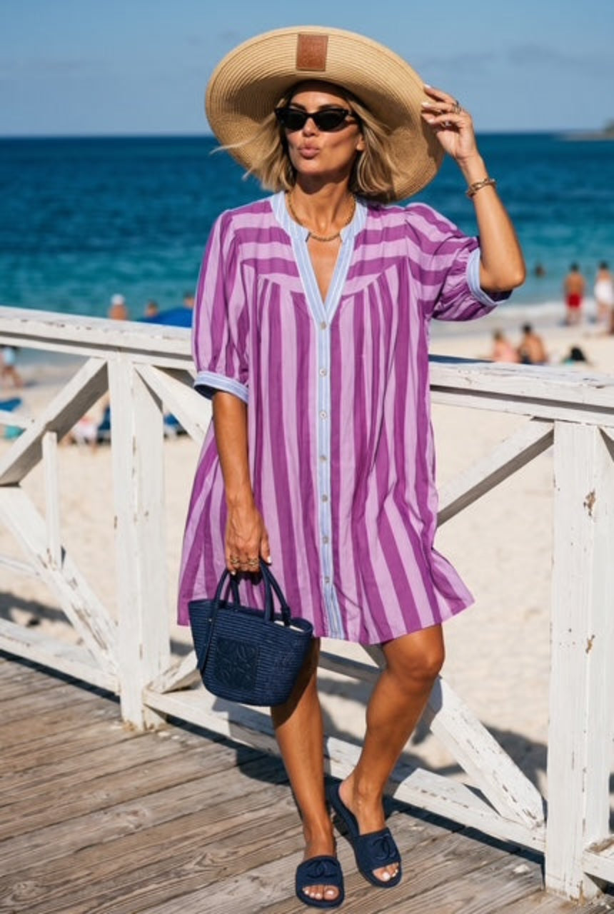 Woman in a purple striped dress and sun hat standing on a wooden deck by the beach.