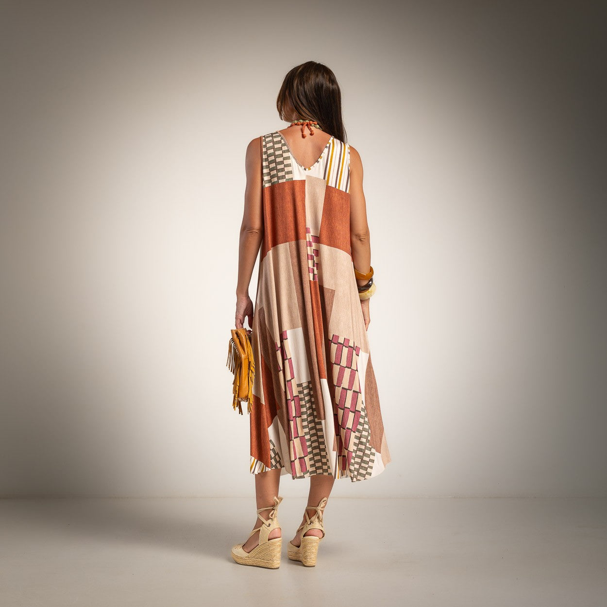 A woman is shown from behind wearing a sleeveless patchwork midi dress with geometric patterns in beige, rust orange, and white. 