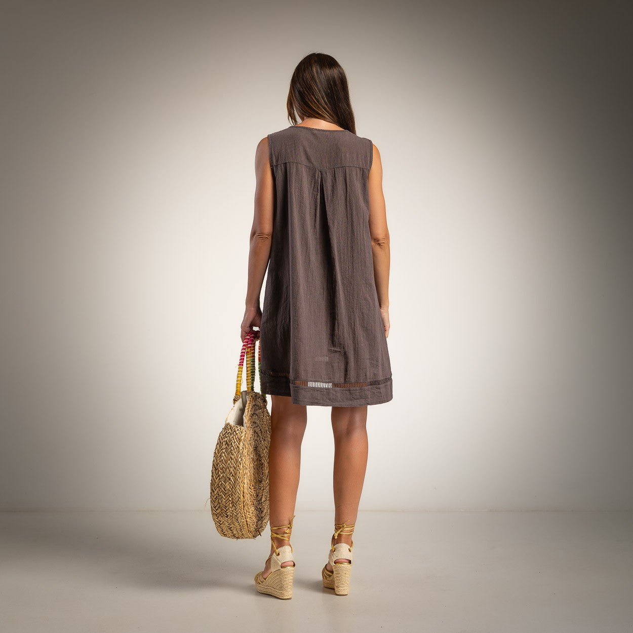 A woman viewed from behind wearing a sleeveless brown dress, holding a woven straw tote bag
