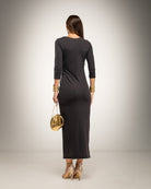 A woman is shown from the back wearing a dark charcoal gray long-sleeve midi dress, holding a small gold handbag and wearing gold high heels 