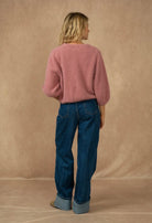 A woman is shown from behind wearing a dusty pink knit cardigan and dark blue jeans with cuffed hems, standing against a textured beige backdrop.