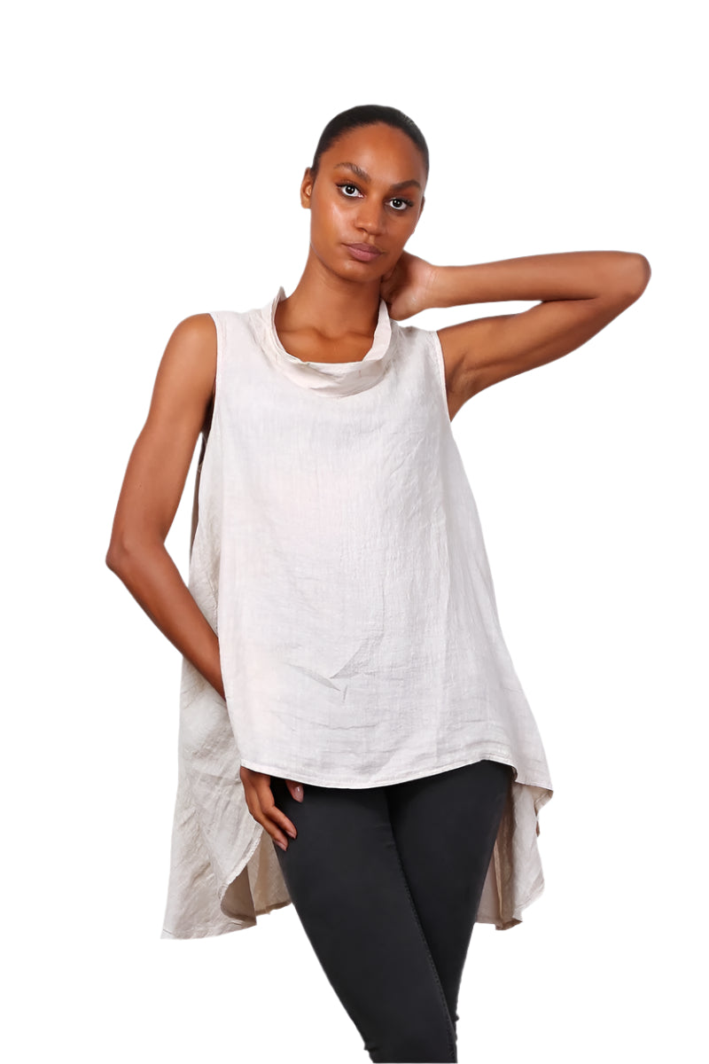 A woman wearing a beige sleeveless cowl neck top with an asymmetrical hem, paired with white trousers.