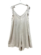 A light beige linen playsuit with tie-shoulder straps, hanging on a white plastic hanger against a wrinkled white fabric backdrop.
