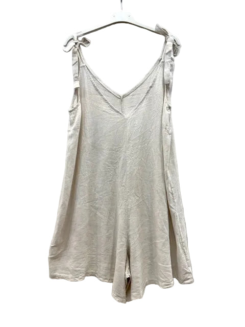 A light beige linen playsuit with tie-shoulder straps, hanging on a white plastic hanger against a wrinkled white fabric backdrop.