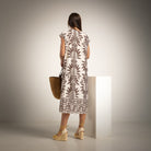 A woman stands with her back to the camera wearing a cream-colored dress with a brown botanical leaf pattern, holding a woven straw bag and standing next to a white display block in a studio setting.
