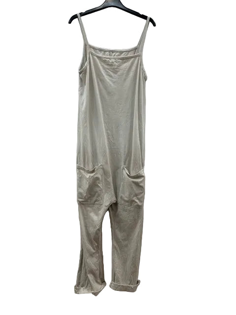 A light beige sleeveless jumpsuit with thin shoulder straps and large front patch pockets, hanging on a black hanger against a wrinkled white fabric backdrop.