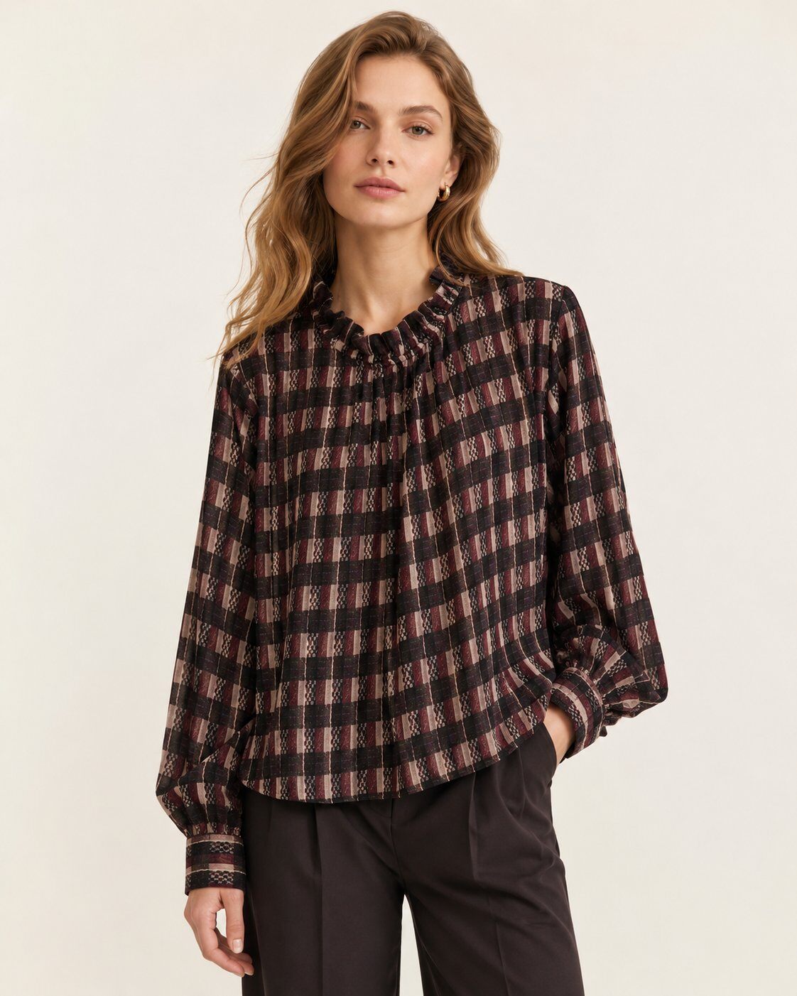 A woman wearing a long-sleeve plaid blouse with a gathered ruffled neckline, paired with dark trousers, against a plain light beige studio background.