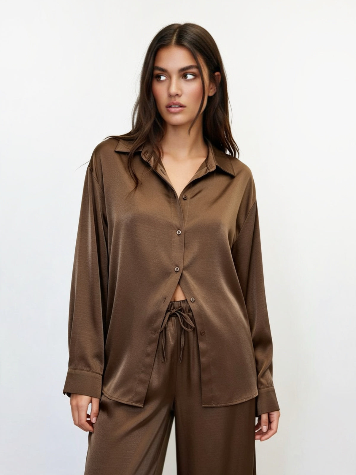 A woman modeling a matching brown satin pajama set, featuring a long-sleeve button-up shirt and coordinating pants, against a plain white studio background.