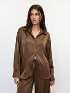 A woman modeling a matching brown satin pajama set, featuring a long-sleeve button-up shirt and coordinating pants, against a plain white studio background.