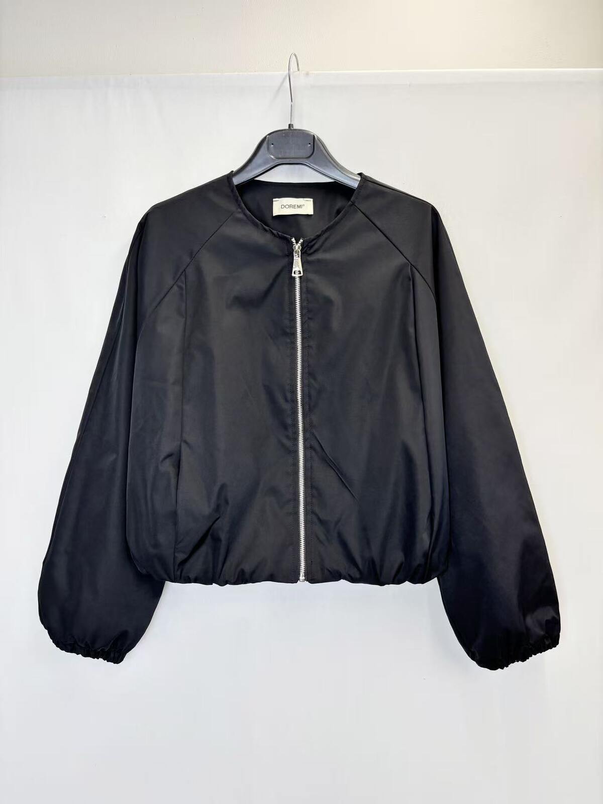 A black lightweight zip-up jacket hanging on a black hanger against a plain white wall. The jacket has a smooth, slightly shiny finish, elasticized cuffs and hem, and a silver zipper.
