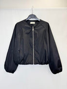 A black lightweight zip-up jacket hanging on a black hanger against a plain white wall. The jacket has a smooth, slightly shiny finish, elasticized cuffs and hem, and a silver zipper.