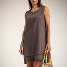 A woman wearing a sleeveless, brown dress, holding a woven straw bag with colorful striped handles 