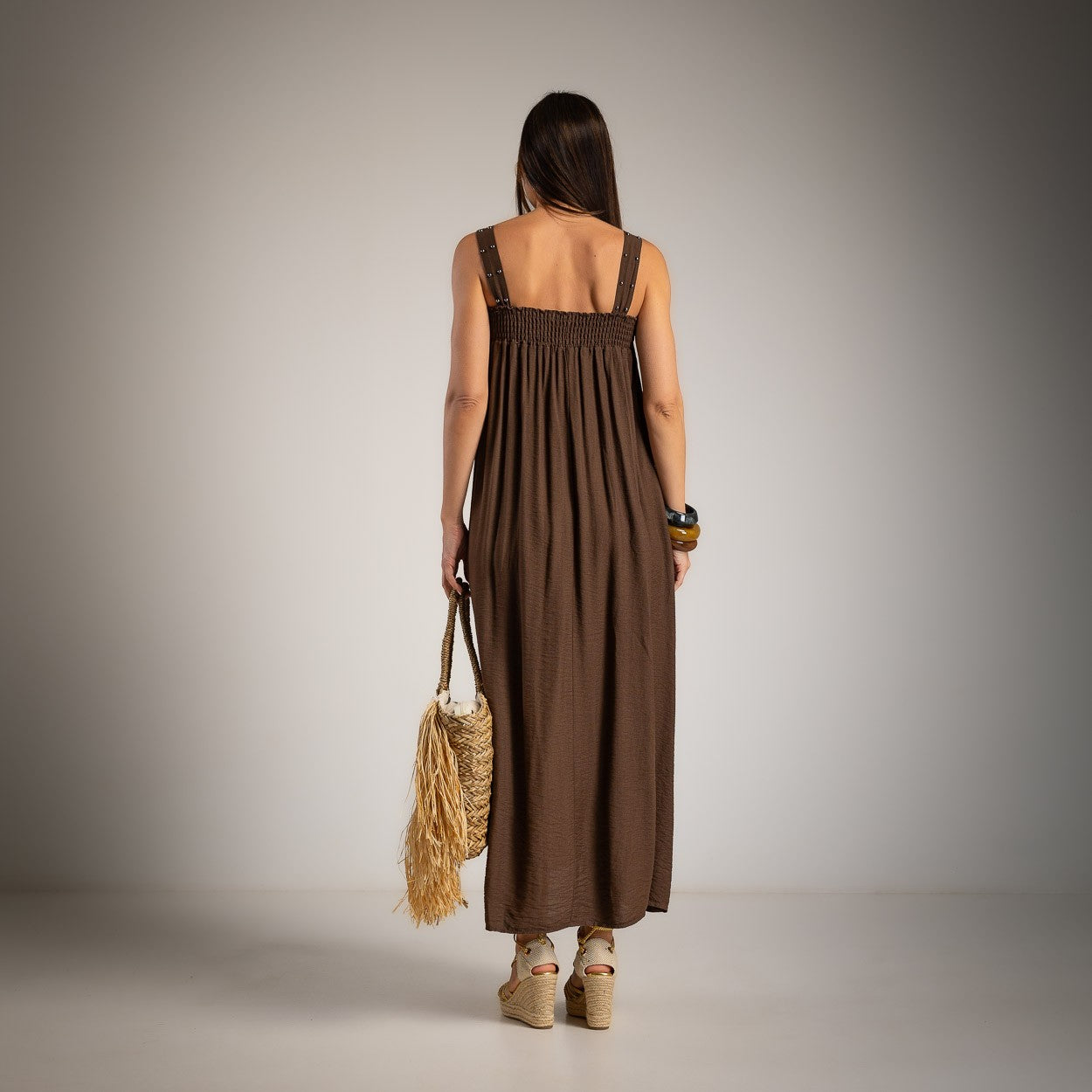 A woman viewed from behind wearing a sleeveless brown maxi dress with a smocked bodice and wide shoulder straps
