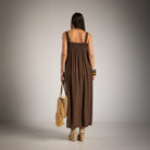 A woman viewed from behind wearing a sleeveless brown maxi dress with a smocked bodice and wide shoulder straps
