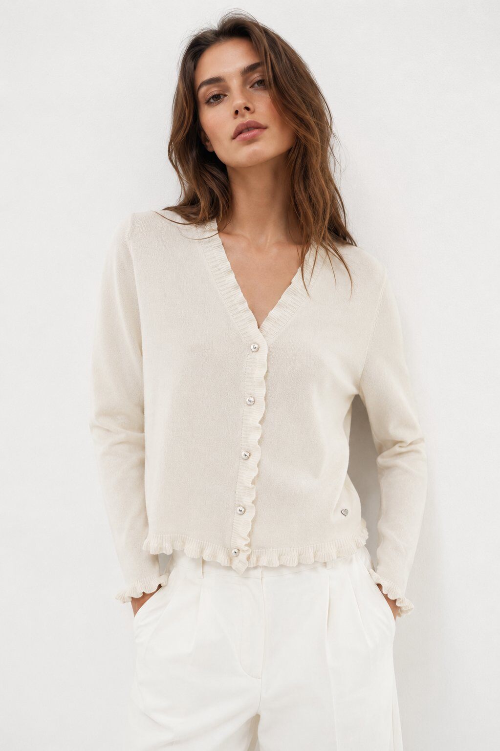 A woman wearing a cream-colored knit cardigan with a scalloped ruffle trim and button front, paired with white trousers, against a plain white background.