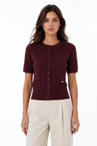 A woman modeling a short-sleeve burgundy knit top with a button-down front, paired with cream-colored trousers, against a plain white background.