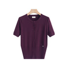 A short-sleeve knit cardigan top in deep plum purple, hanging on a light wood hanger against a clean white background. The cardigan features a round neckline, a front button closure, and a small silver heart detail on the lower right side.