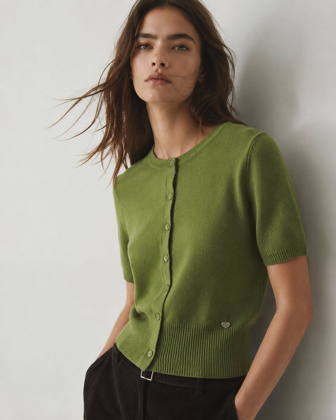 A woman wearing a short-sleeve knit cardigan in olive green with a button-front closure and ribbed hem, paired with dark pants, posed against a plain light gray wall.