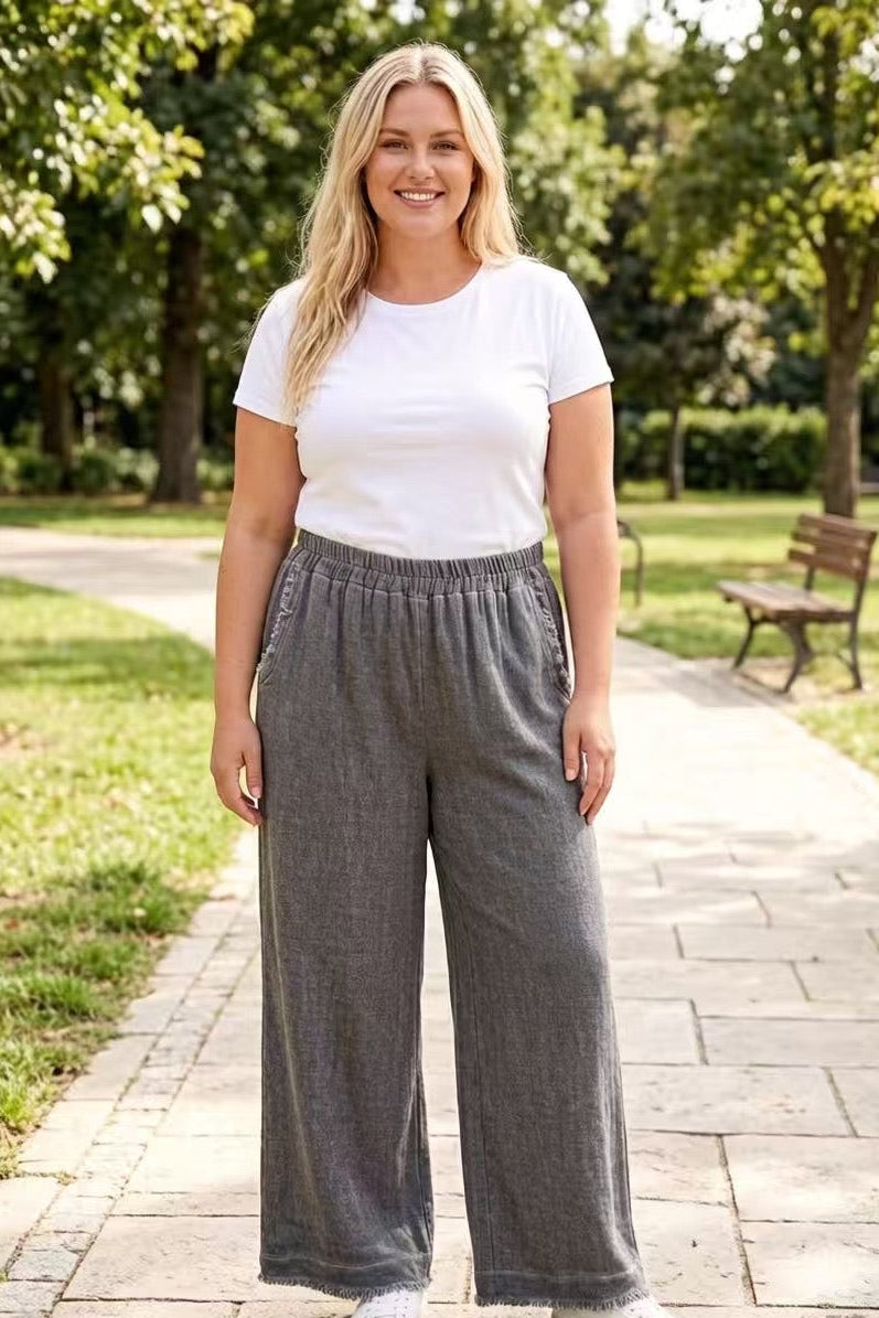 Woman wearing a white t-shirt and charcoal-grey-wide-leg pants standing on a paved path with greenery in the background.