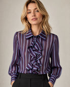 A woman wearing a sheer long-sleeve blouse with vertical purple, blue, and white stripes and a large ruffled tie-neck detail, paired with black trousers, against a neutral beige background.