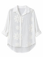 A white long-sleeve button-up blouse with a pointed collar, featuring a textured gingham pattern and large floral applique details, shown against a plain white background.