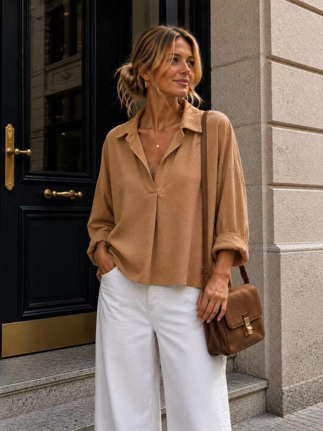 A woman stands on stone steps in front of a black door, wearing a camel shirt, white wide-leg pants, and carrying a brown leather shoulder bag.