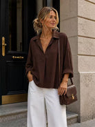 A woman stands on stone steps in front of a black door, wearing an oversized dark brown blouse with white wide-leg pants and carrying a brown leather crossbody bag.