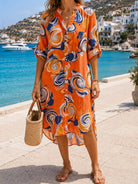 A woman wearing a vibrant orange button-up shirt dress with a blue, white, and yellow swirl pattern, standing on a sunny seaside promenade with a woven straw bag.