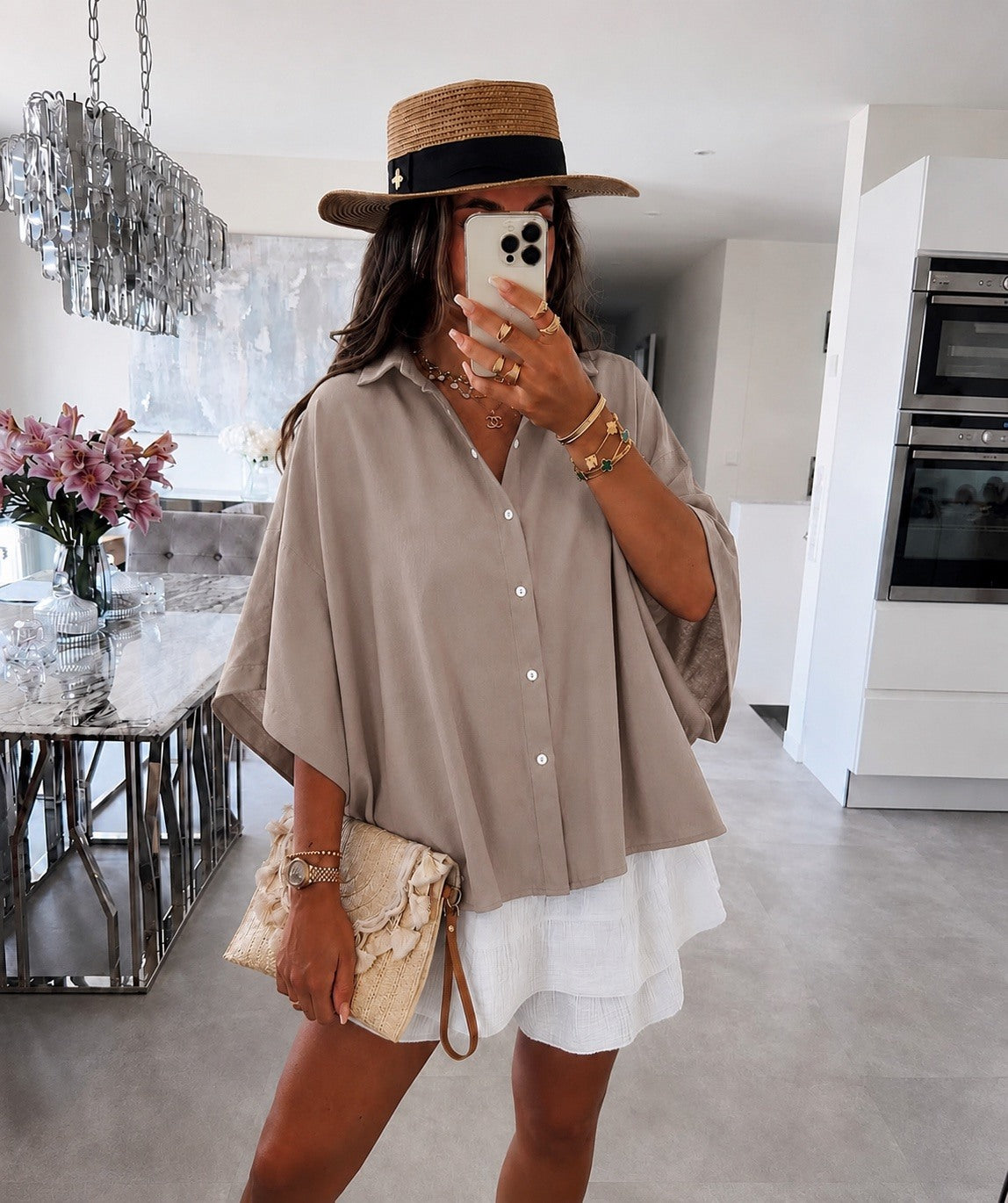 A woman takes a mirror selfie in a modern white kitchen, wearing a beige oversized short-sleeve button-up shirt, a white ruffled mini skirt, a straw hat with a black band, and holding a beige clutch bag.