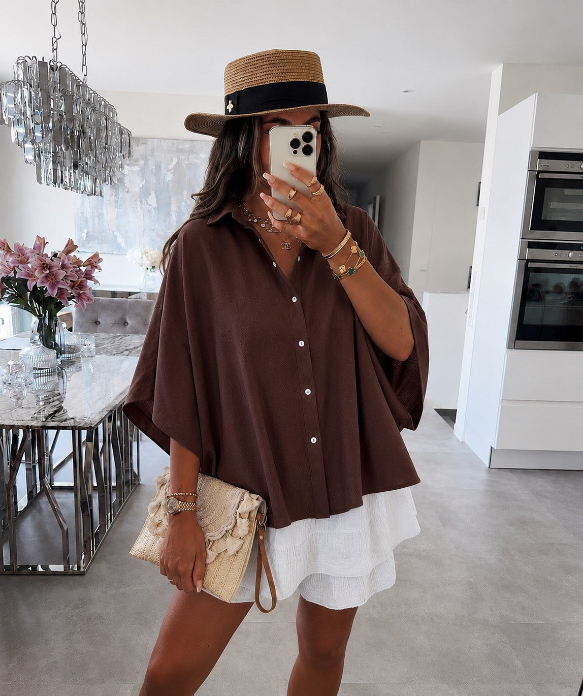 A woman takes a mirror selfie in a modern kitchen setting, wearing a brown short-sleeve button-up shirt