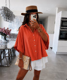 A woman takes a mirror selfie in a modern kitchen setting, wearing a vibrant orange oversized short-sleeve button-up shirt