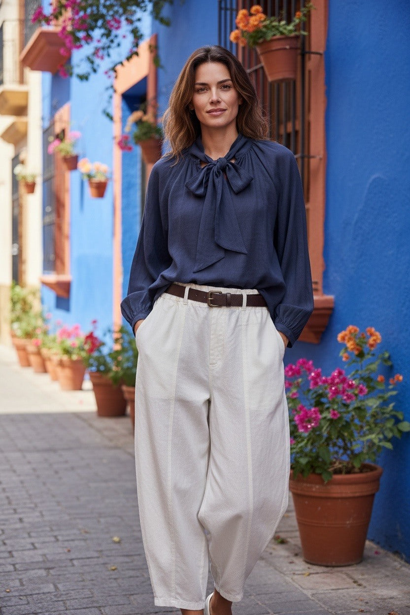 A woman stands on a cobblestone street in front of a bright blue building, wearing a navy blue long-sleeve blouse with a large bow tie neckline and white wide-leg trousers.