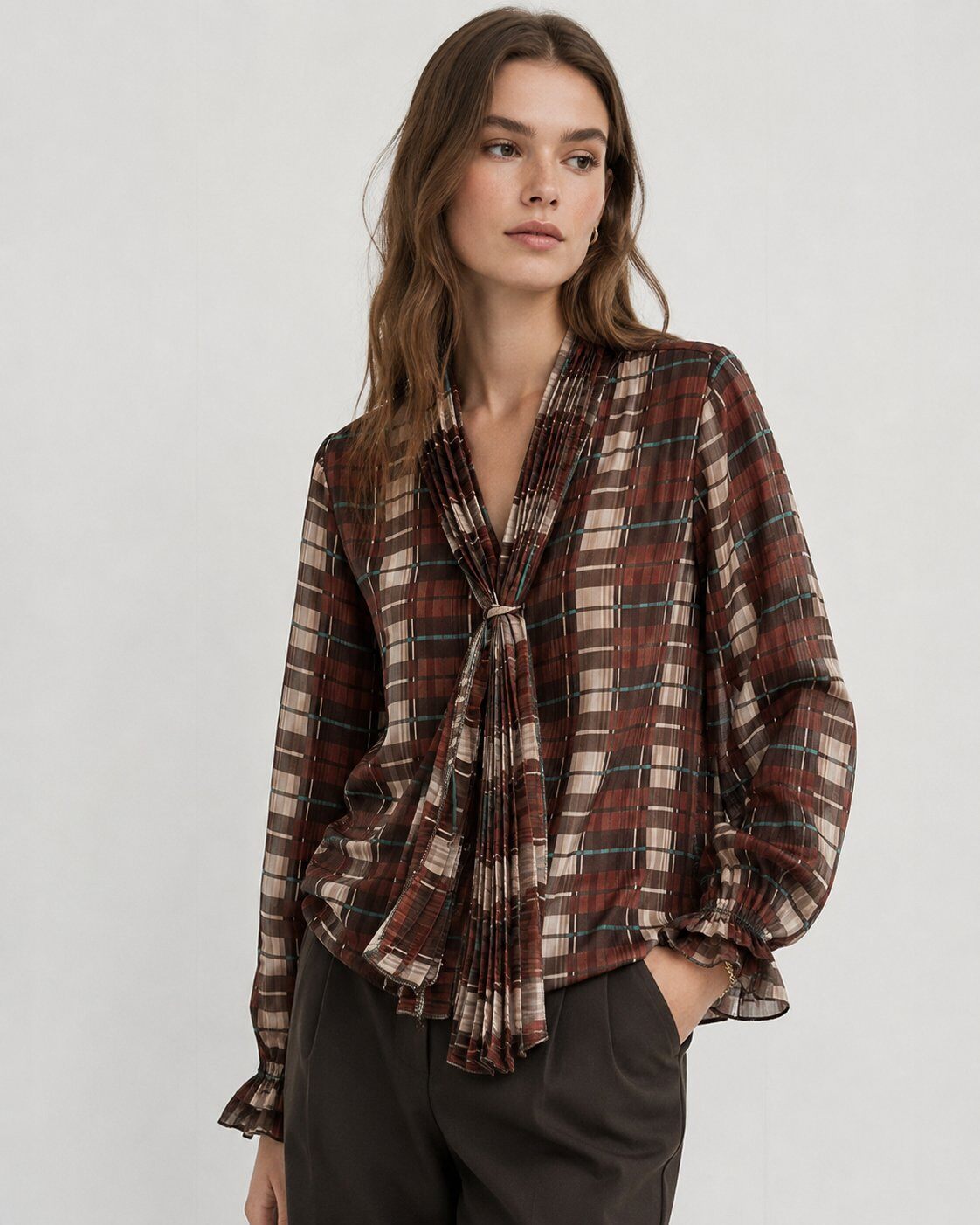 A woman modeling a long-sleeve plaid blouse with a gathered, tie-front neckline and ruffled cuffs, paired with dark trousers against a light gray studio background.