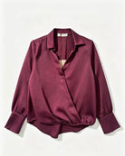 A long-sleeve satin blouse in deep burgundy, featuring a pointed collar, a partial button placket, and gathered cuffs, laid flat against a light off-white background.