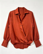A rust-orange long-sleeve blouse with a pointed collar, button front, and gathered cuffs, laid flat against a light off-white background.
