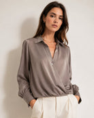 A woman wearing a taupe satin long-sleeve wrap blouse and cream trousers, standing against a neutral light gray wall.