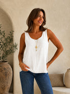 A woman wearing a white sleeveless top and blue jeans, standing in a warm, neutral-toned interior setting with a textured wall, potted plant, and cushions.