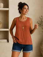 A woman wearing a sleeveless rust-orange linen top and dark blue denim shorts, accessorized with a gold key necklace, standing in a warm, neutral-toned room.