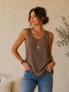 A woman wearing a brown sleeveless top and blue jeans, standing against a textured beige wall with a decorative plant in the background.