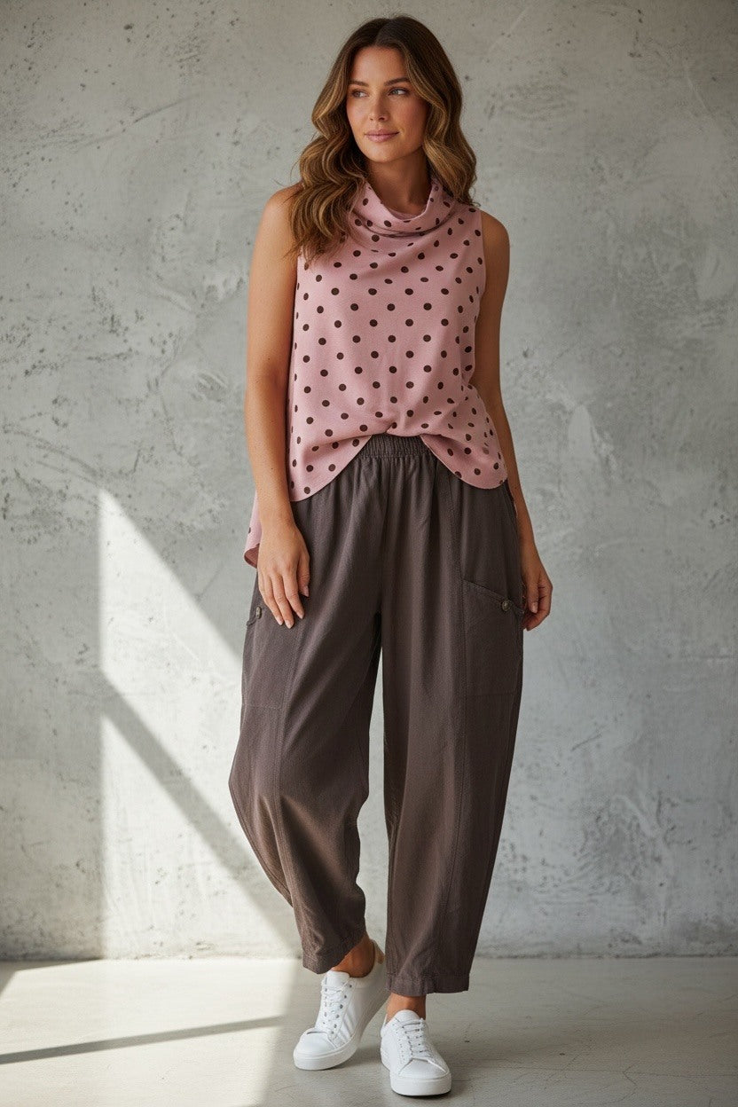 A woman modeling a sleeveless dusty pink top with black polka dots and dark brown wide-leg pants, standing against a textured gray concrete wall.