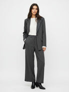 A woman modeling a dark gray pinstripe blazer and matching wide-leg trousers, styled with a white top and black ankle boots, against a plain white studio background.