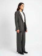 A woman modeling a dark gray pinstripe blazer and matching wide-leg trousers over a white top and black shoes, photographed against a light gray studio background.
