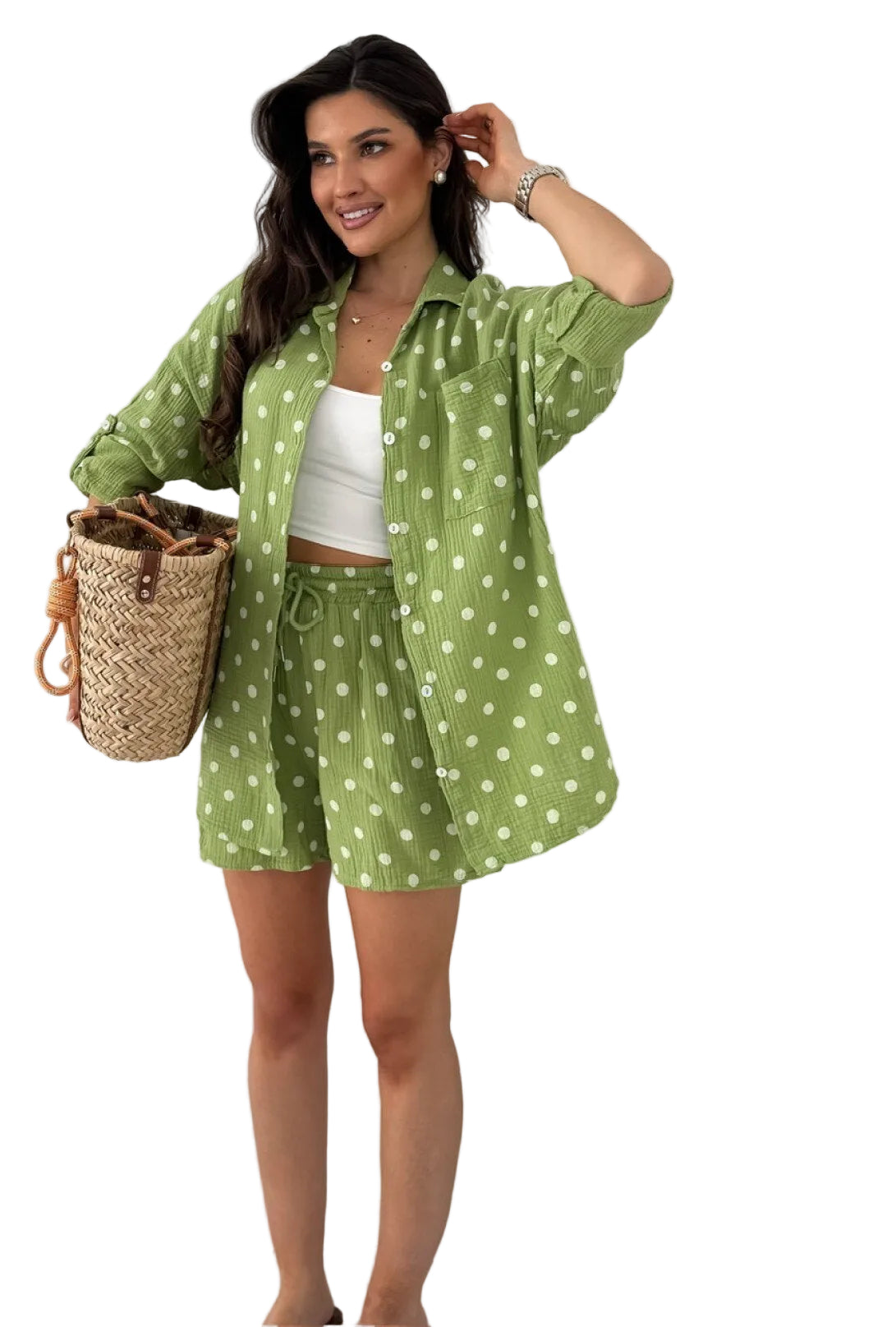 A woman models a matching green and white polka dot lounge set consisting of an oversized button-down shirt and shorts, styled with a white crop top, a woven straw bag, and standing next to a potted cactus.