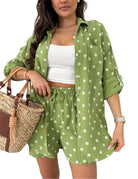 A woman models a matching green and white polka dot two-piece set consisting of a long-sleeve button-up shirt and drawstring shorts, styled with a white crop top and a woven straw handbag.