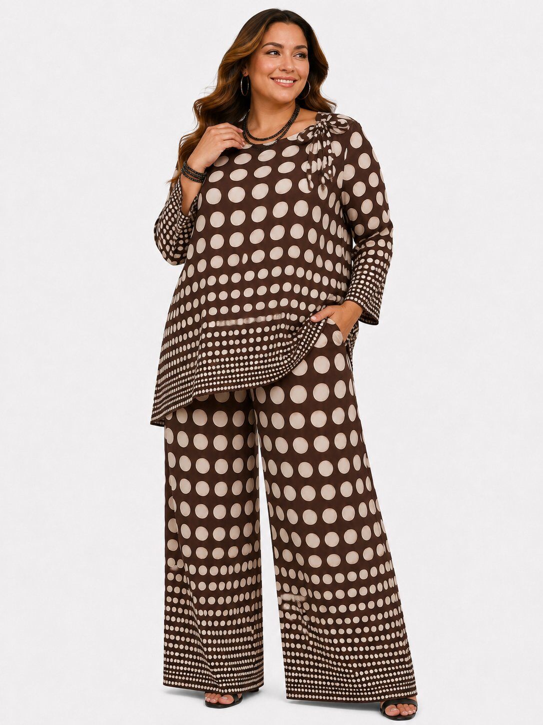 A woman modeling a matching two-piece outfit featuring a brown and beige polka dot pattern, consisting of a long-sleeve ruffled blouse and wide-leg pants, against a plain light gray background.