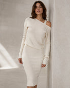 A woman models a ribbed knit two-piece set consisting of a long-sleeve asymmetrical off-the-shoulder crop top and a matching ribbed midi skirt in a warm off-white color, set against a textured concrete wall.