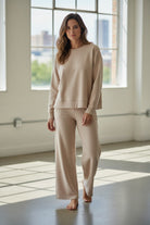 Woman wearing a beige outfit standing in a bright room with large windows.
