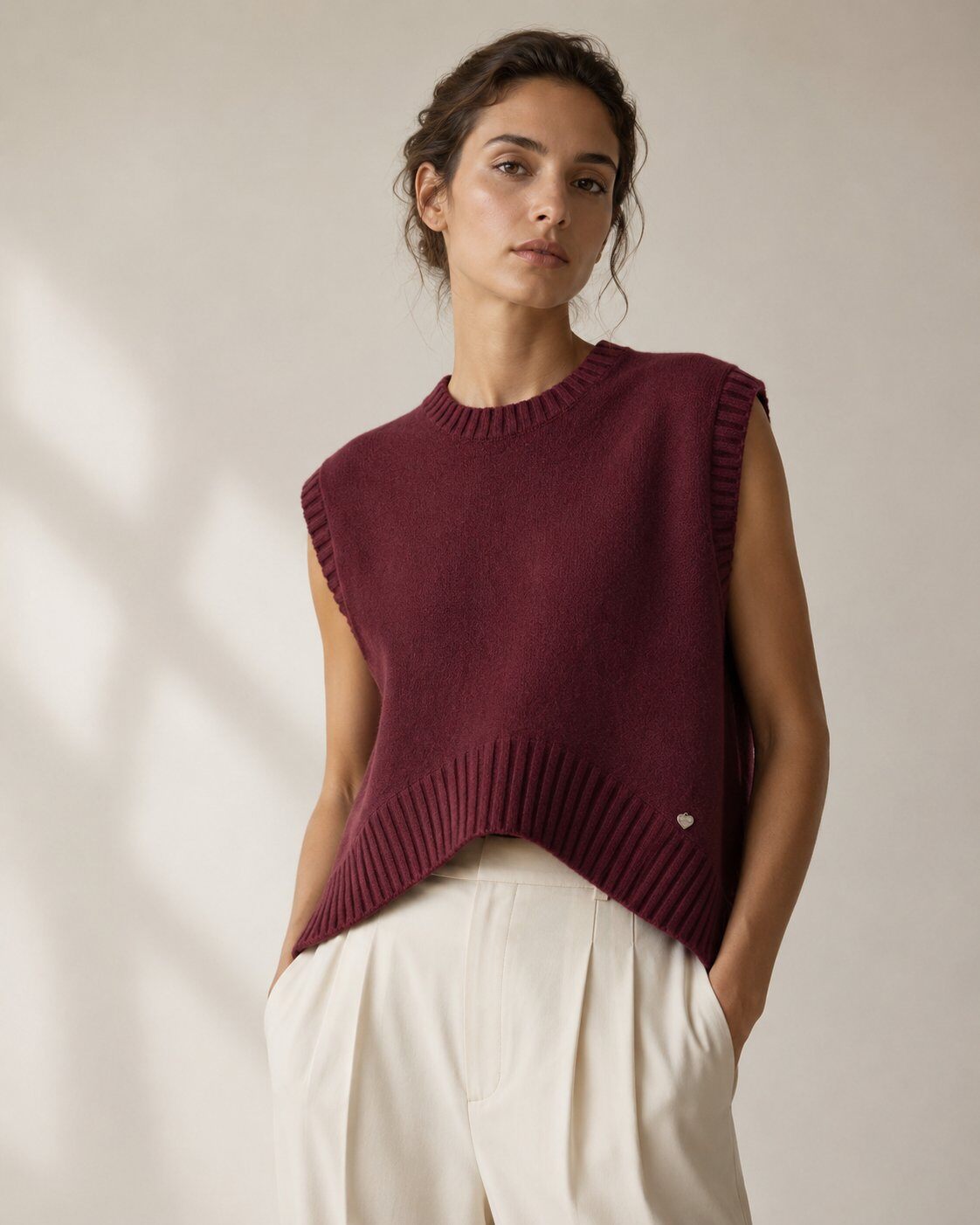 A woman wearing a sleeveless ribbed knit sweater vest in deep burgundy, paired with cream-colored trousers, standing against a warm beige wall with soft window shadows.